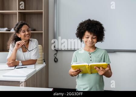 smiling african american teacher looking at schoolboy reading book in classroom Stock Photo