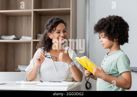 african american boy reading book to smiling teacher during lesson Stock Photo