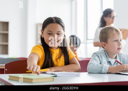 cheerful asian schoolgirl reaching book while sitting at desk near classmate Stock Photo