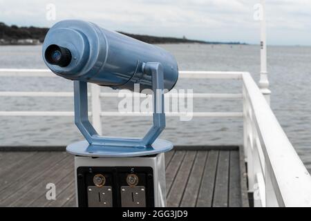 Blue telescope mounted on a stand on wooden ocean pier with white railing. Cloudy summer day on the pier of Jurata, Poland. Stock Photo
