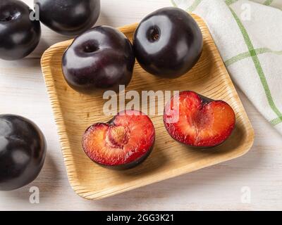 Black plums whole and cut in half on a square wooden plate over table. Ready to eat fresh ripe plums with red juicy pulp. Close-up of tasty sweet frui Stock Photo