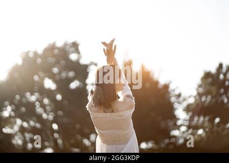Unity with nature. Beautiful native american shaman woman in white dress and feather in hair, performing ritual, standing with arms raised to the sky and looking up. Tribal concept Stock Photo