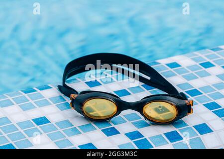 Black professional swimming goggles with water drops on yellow lenses on a blue and white tiles of pool floor. Stock Photo