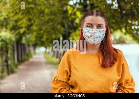 Happy Caucasian millennial woman wearing protective mask outdoor in city park Stock Photo