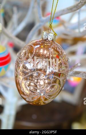 Shiny glittery golden Eastern ornament hangs by a gold cord from white branches with other colorful egg ornaments blurred behind Stock Photo