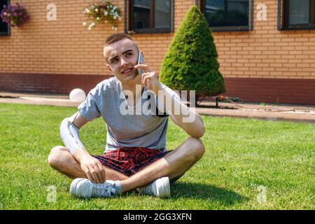 Man with amputated arm and prosthesis talking on the phone sitting on the lawn Stock Photo