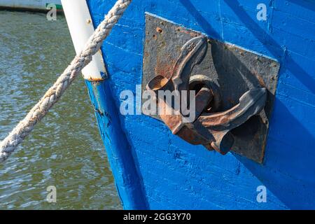 Old rusty ship anchor in up position on moored blue small boat. Close up Stock Photo