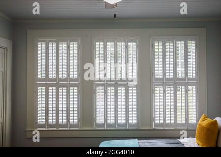 Three large windows with cream colored plantation shutters that are closed on the interior of a cottage style house Stock Photo