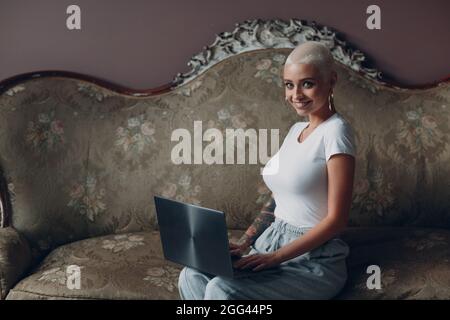 Millenial european young woman with short blonde hairstyle sitting and smiling with laptop. Working on home office freelance concept. Stock Photo