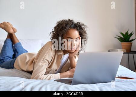 African American female student sitting on bed at home typing on laptop. Stock Photo