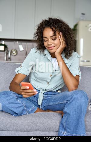 Serene mixed race relaxed student girl sit on sofa at home holding cellphone. Stock Photo