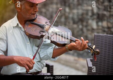 Street old musician sitting on a chair with a cap on his head wearing striped t-shirt, playing violin for all people who pass by him and listen to his Stock Photo