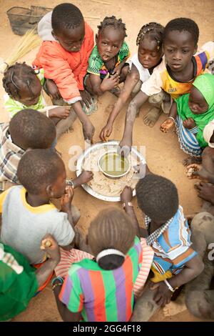 A group of children eat around a communal dish of millet paste and baobab leaf stew, likely their only meal of the day. This extended family has been displaced due to violence and insecurity and now faces hunger and food shortages. Kossi Province, Burkina Faso, West Africa. Stock Photo