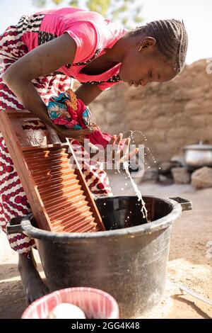 A girl washes clothes with a washboard and basin outside her home in Ségou Region, Mali, West Africa. Stock Photo