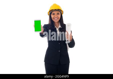 portrait of mixed race asian/caucasian engineer woman with helmet holding blueprints and showing green screen mobile phone isolated white background Stock Photo