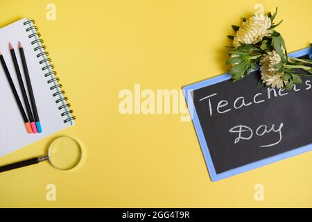 Asters flowers lying down on a chalkboard with text Teachers Day , and a magnifier loupe next to colored pencils on a blank sheets of a notepad isolat Stock Photo