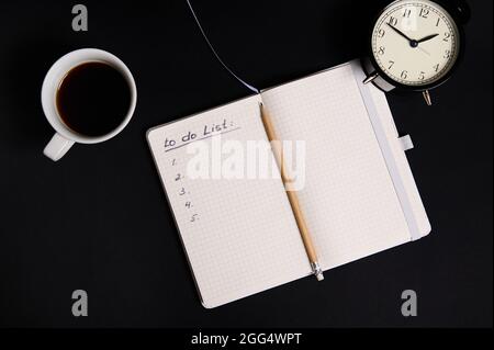 Flat lay of an opened organizer, notepad with list to do on blank white sheets, cup of coffee and alarm clock on black background with copy space. Bus Stock Photo