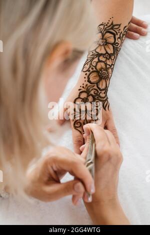 Mehndi artist drawing floral henna tattoo on woman hand in beauty salon. Master applying traditional mehendi pattern for bride before wedding. Stock Photo