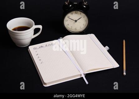 Opened organizer, notepad with list to do on blank white sheets, wooden pencil, cup of coffee and alarm clock on black background with copy space. Bus Stock Photo