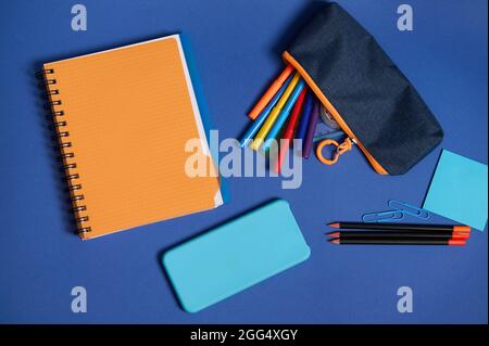 Top view of school office supplies and a smartphone lying screen down on a blue background . Flat lay composition of stationery in two contrasting col Stock Photo