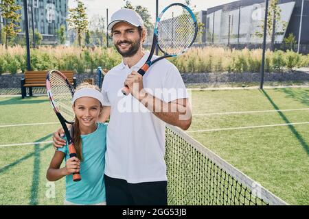 Portrait of father tennis player and little daughter in sportswear with rackets in hands on tennis court outdoor near net. sporty family playing tenni Stock Photo