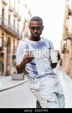 african man standing on the streets of Barcelona. He is using his smart phone and holds a white reusable cup of coffee Stock Photo