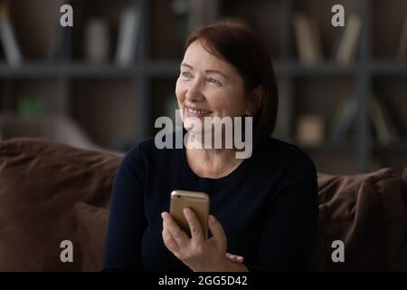 Dreamy smiling mature woman holding smartphone in hands. Stock Photo