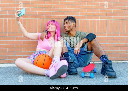 alternative diverse couple hanging out together chilling and smiling sitting against background wall holding smartphone. happy interracial friends Stock Photo