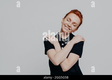 Happy red-haired woman with closed eyes dressed casually hugging herself gently and tilting head Stock Photo