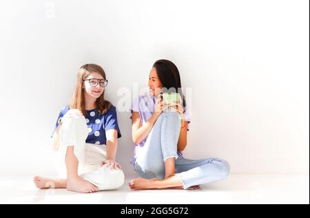 Two happy casual women relaxing with hot drink in new home. Young girls chatting talking about funny things gossip laughing in light apartment. Female Stock Photo