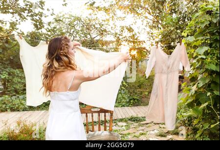 Young woman hanging laundry outdoors. Cute girl in dress washing white clothes in metal basin in backyard, hanging laundry on clothesline and leaving Stock Photo