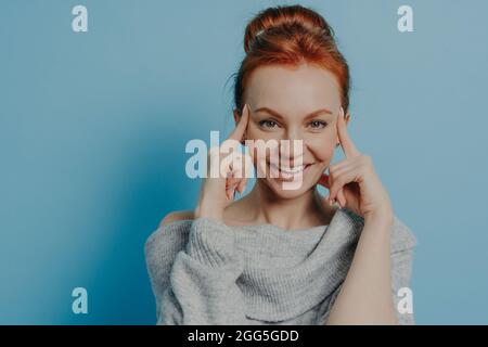 Young positive red haired woman with fingers on temples standing isolated on blue background Stock Photo