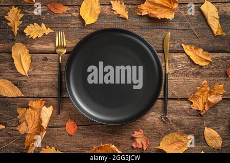 A black plate and cutlery among autumn leaves on a wooden background. Stock Photo