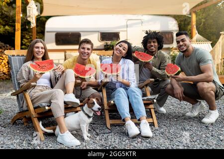 Happy diverse friends eating watermelon near RV, sitting in lounge chairs and enjoying autumn camping trip with pet dog Stock Photo