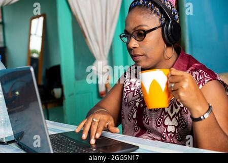 Attractive young woman sitting inside the house, working on laptop, wearing headphones, drinking coffee Stock Photo