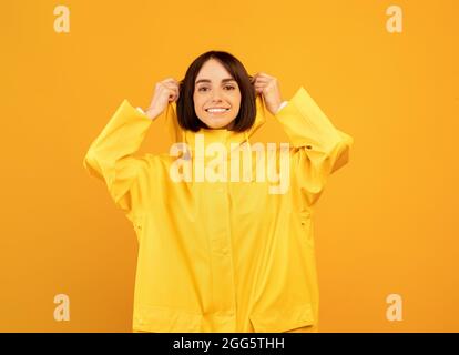 Happy woman in yellow waterproof raincoat putting on hood, standing isolated on yellow background Stock Photo