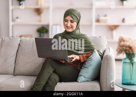 Cheerful Mature Muslim Female Using Laptop Sitting On Couch Indoor Stock Photo