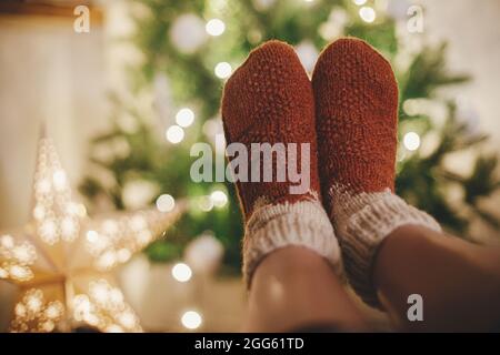 Woman feet in cozy woolen socks on background of christmas tree and star in lights in festive evening room. Cozy winter moments at home. Stylish warm