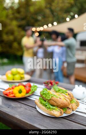 Sandwiches and vegetables, picnic foods on table outdoors, group of young friends toasting with beer on background Stock Photo