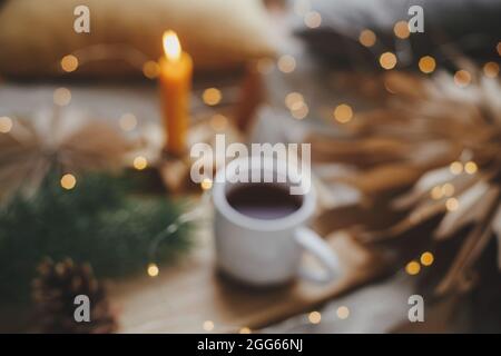 Blurred image of warm cup of tea with christmas lights, stars, pine trees, candle on bed in scandinavian evening room. Winter hygge abstract backgroun Stock Photo