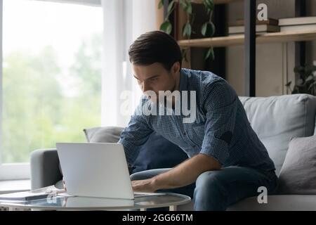 Serious millennial man using laptop, typing, sitting on couch Stock Photo