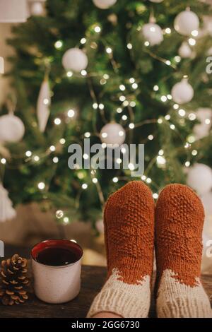 Cozy winter moments at home. Woman feet in cozy woolen socks and cup of warm tea on background of christmas tree in lights in festive evening room. St