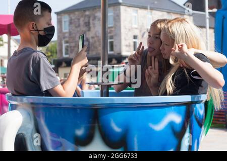 Teenagers on the teacup ride at the summer fayre, Helensburgh, Scotland using mobile phones and wearing a mask. Stock Photo