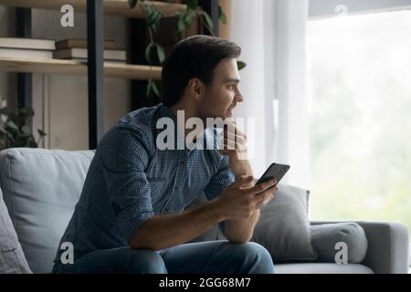 Happy thoughtful millennial man holding mobile phone Stock Photo