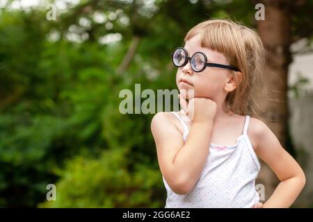 Young clever intelligent school age child, little girl in big quirky glasses thinking, wondering, deep in thought, copy space, outdoor portrait, face Stock Photo