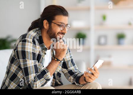 Cheerful long-haired middle-eastern guy hipster sitting on couch, using smartphone Stock Photo