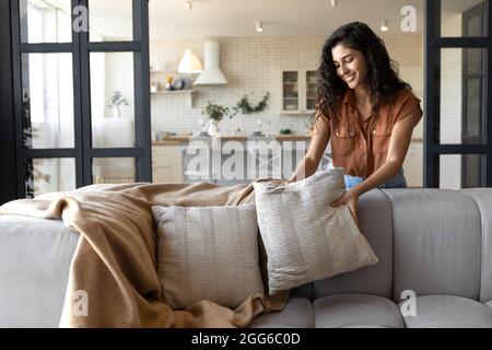 Lovely young woman putting soft pillows and plaid on comfy sofa, making her home cozy and warm, copy space Stock Photo
