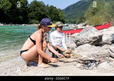 Mother and son toasting marshmallows on a camp fire on riverbank of Soca river, canoe behind, Slovenia Stock Photo