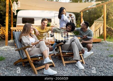 Young multiracial friends playing guitar and singing, holding dog, camping together, having fun autumn vacation outdoors Stock Photo