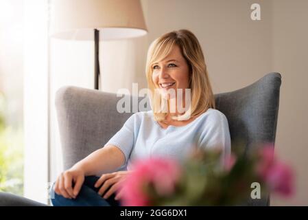 Smiling blond haired woman relaxing at the airmchair at home. Stock Photo